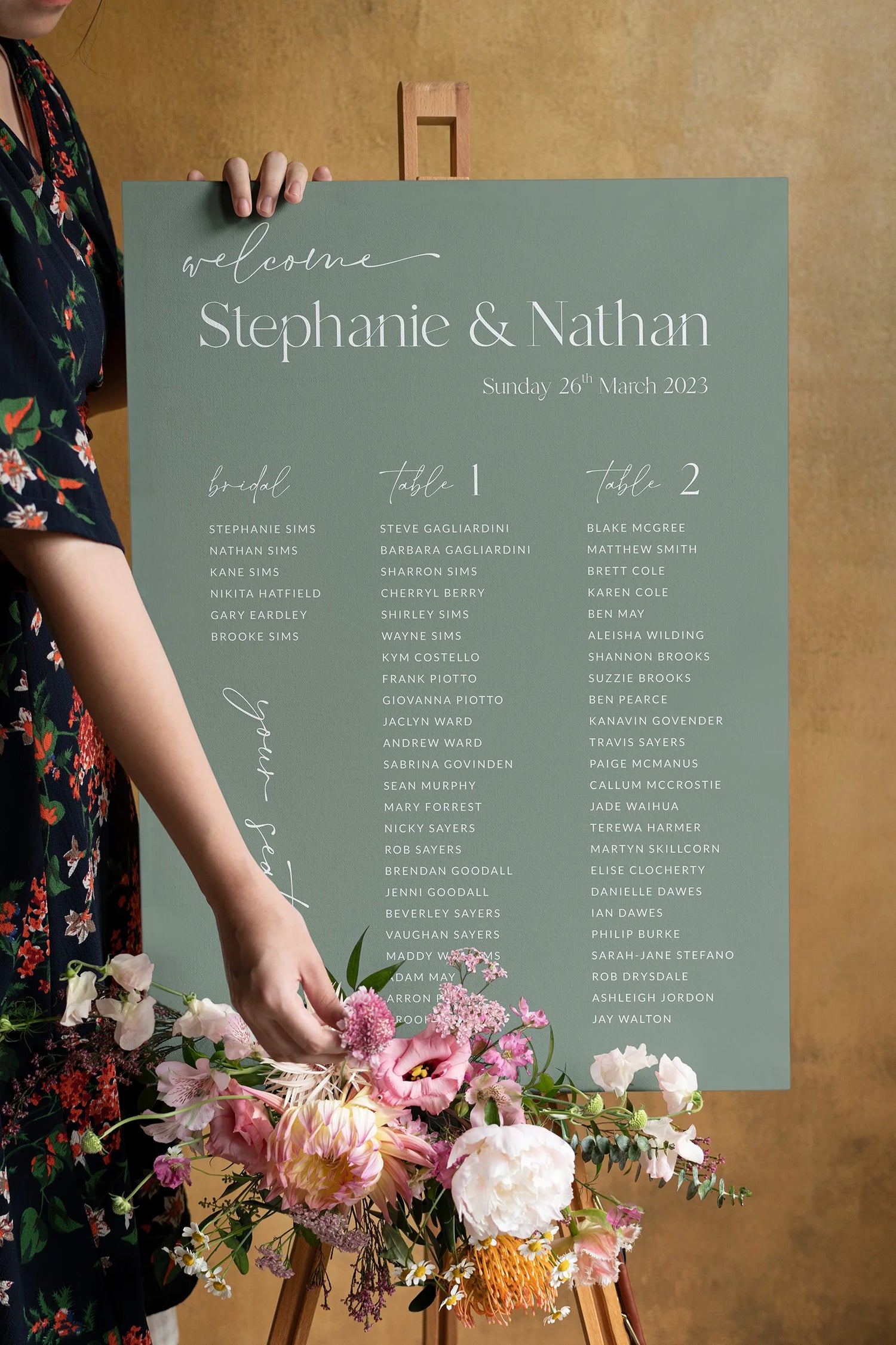 Modern wedding seating chart on a sage green background with white calligraphy, elegantly displayed on a wooden easel and accented with soft pastel flowers