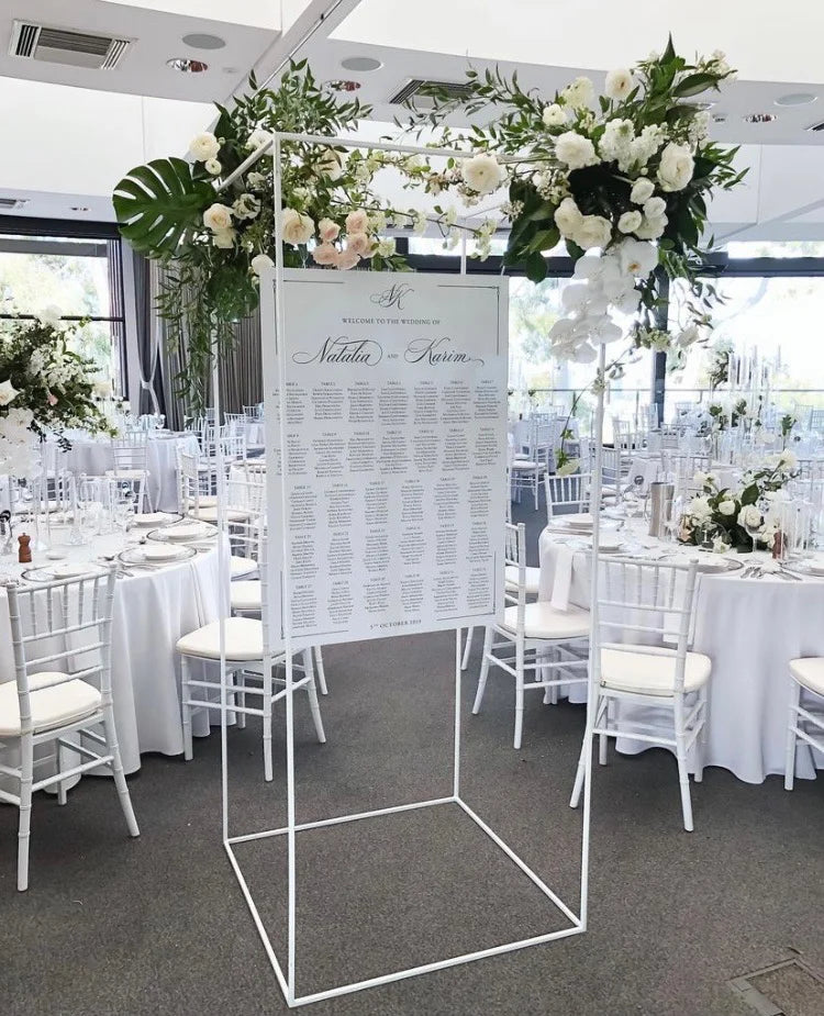 Wedding seating chart displayed on a modern white metal stand, adorned with lush white flowers and green foliage, set in an elegant reception venue with round tables and white chairs.