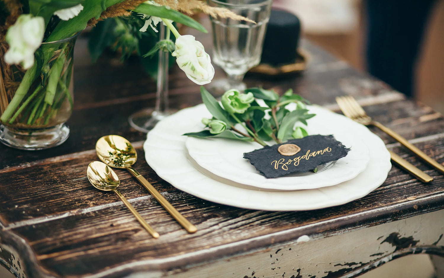 Vintage-inspired wedding table setting with gold cutlery, a white plate, and a black slate place card featuring gold calligraphy and greenery.