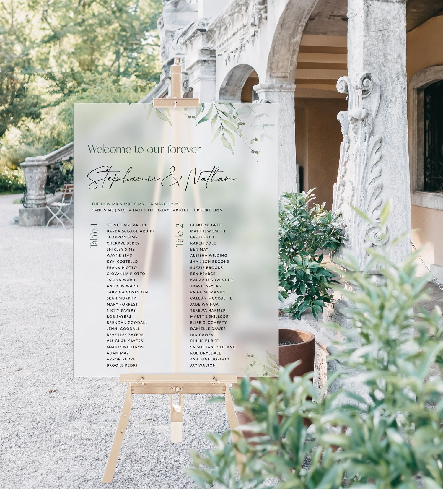  An elegant acrylic wedding seating chart for Stephanie and Nathan, displayed on a wooden easel outdoors. The sign features a soft green botanical design with table assignments listed in two columns. A classical stone building and lush greenery provide a scenic backdrop.