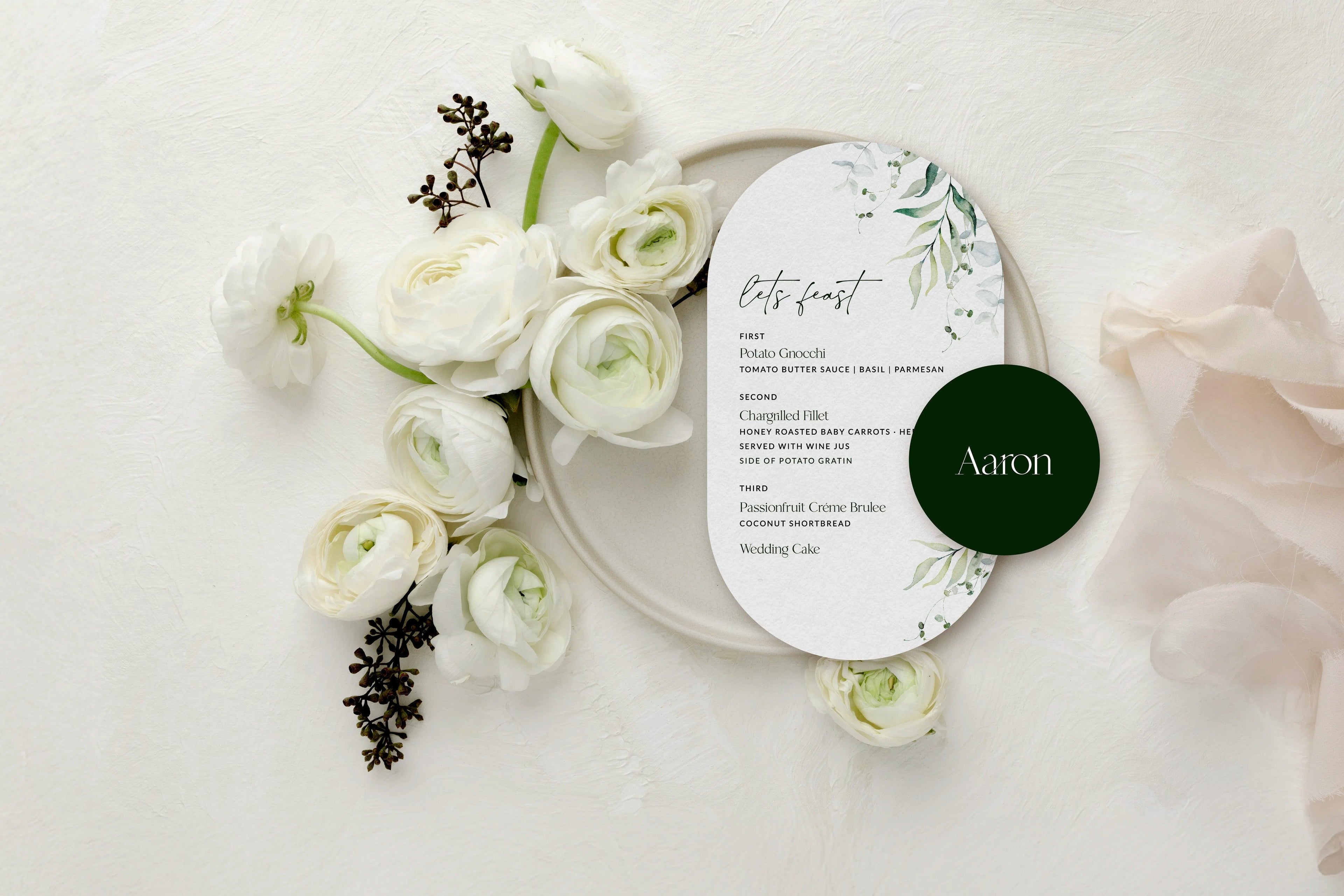 Elegant wedding menu card with botanical accents on a beige plate, surrounded by white ranunculus flowers and greenery, featuring a dark green name tag reading 'Aaron'.