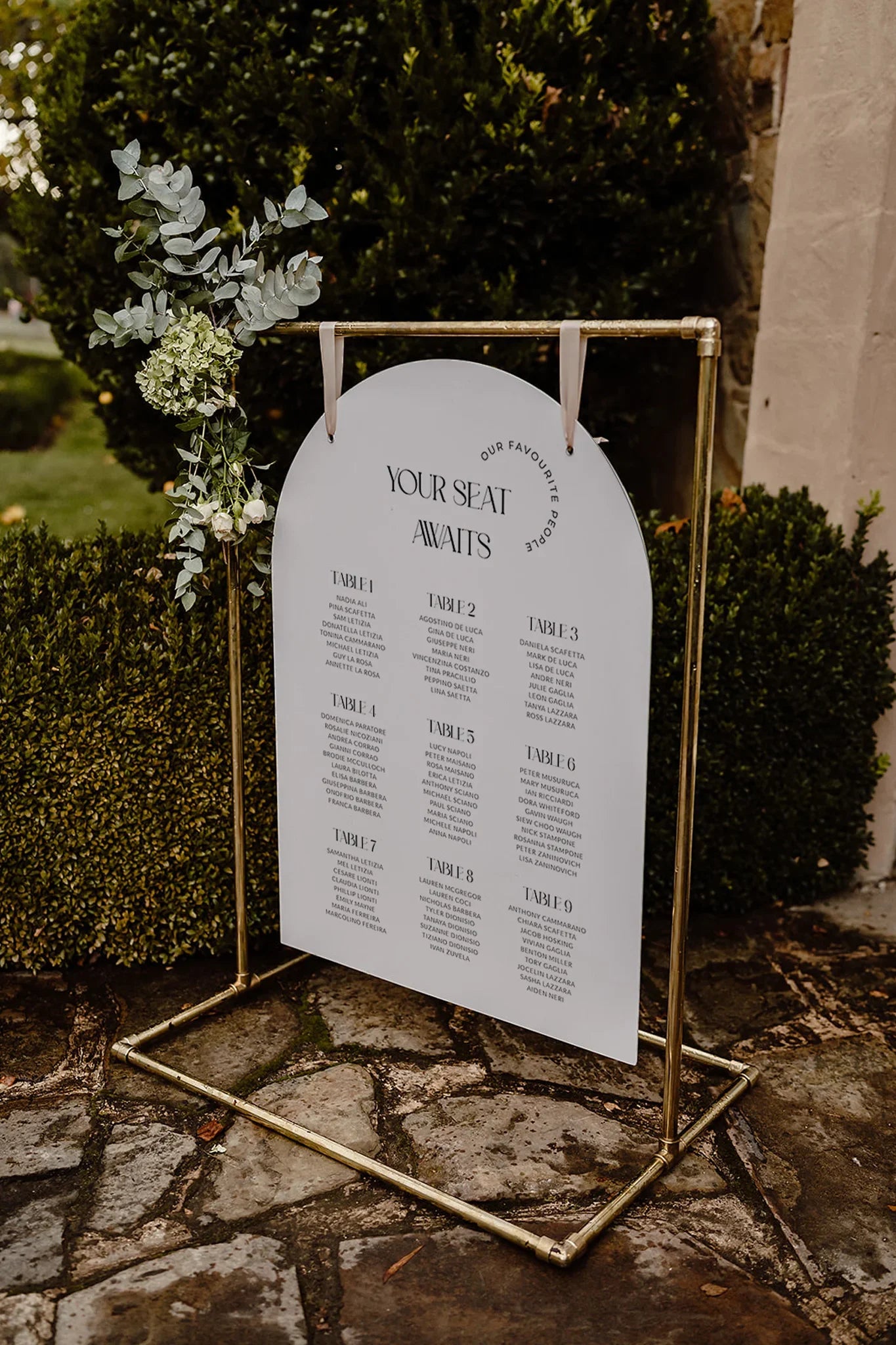 Stylish wedding seating plan displayed on a gold stand, featuring an arch-shaped sign with guest table assignments and decorated with eucalyptus and white flowers