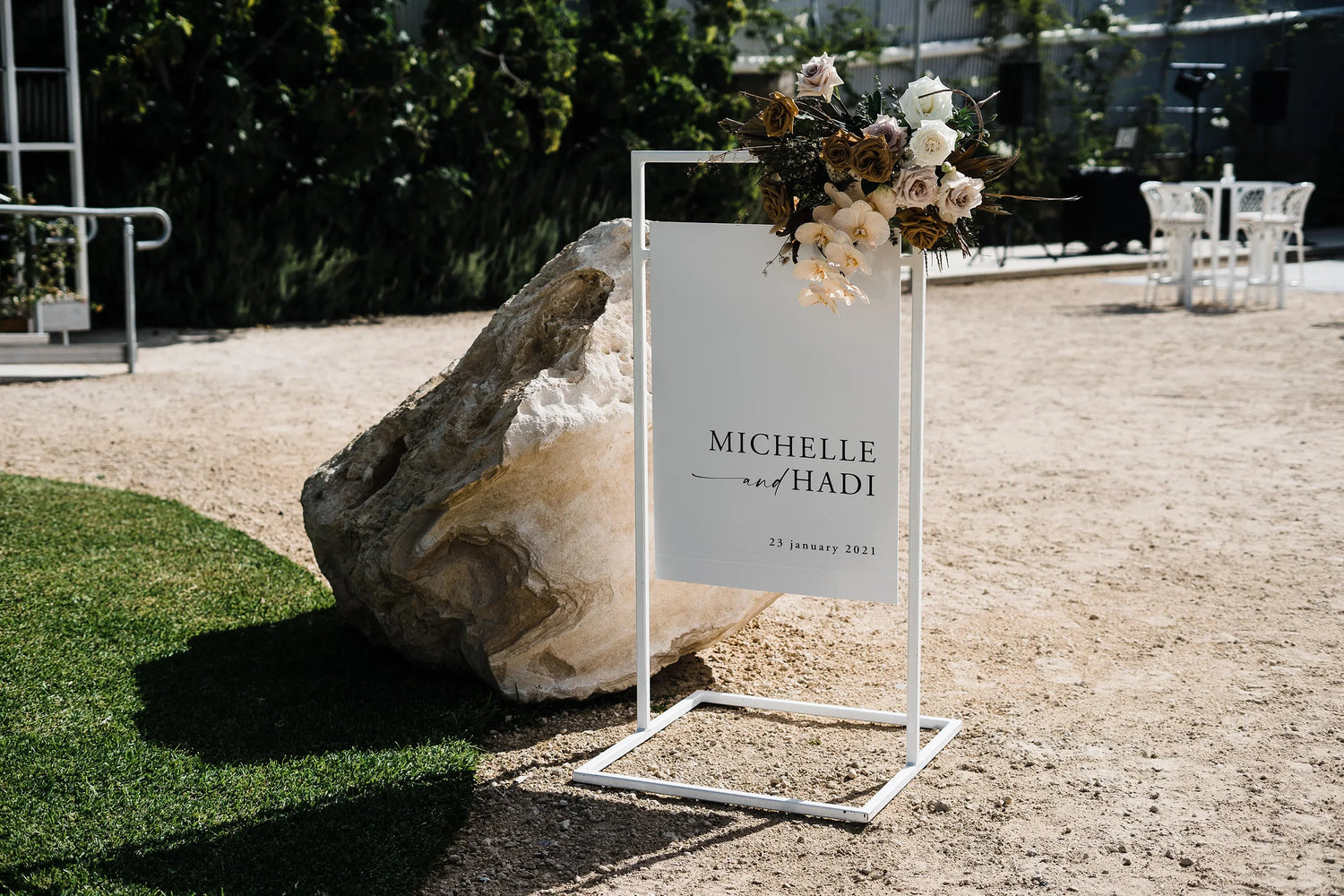 Elegant white welcome sign for Michelle and Hadi’s wedding, displayed on a sleek white stand, adorned with floral arrangements in neutral tones against a natural outdoor backdrop.