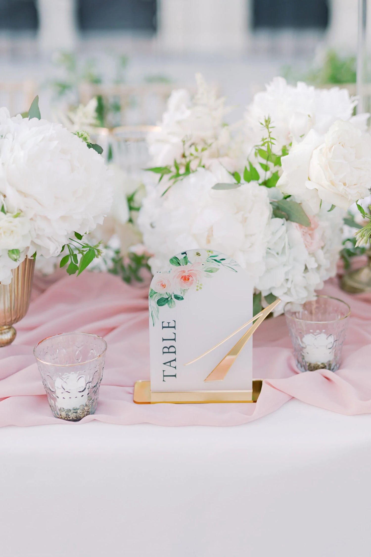 Modern acrylic table number with floral illustrations and gold accents, displayed on a blush-pink tablecloth surrounded by lush white flowers and glass votive candles.