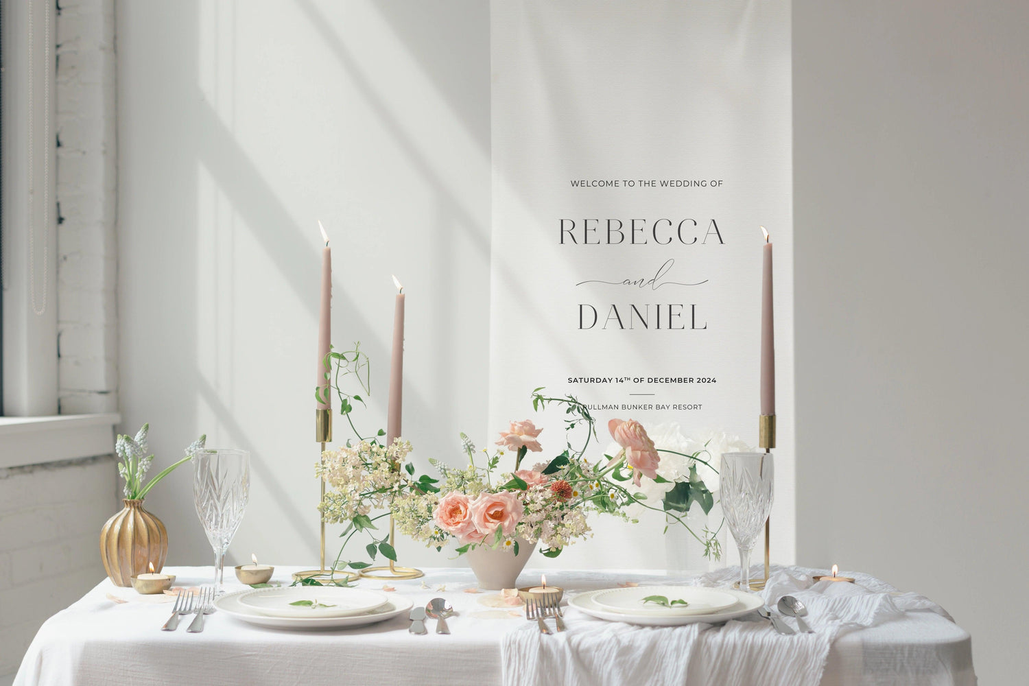 Wedding table setting featuring a linen welcome sign with 'Rebecca and Daniel' in elegant typography, flanked by blush pink candles, floral arrangements in soft pastel tones, and tableware on a white tablecloth.