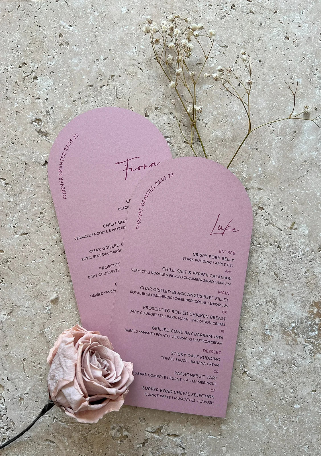 Two blush-pink arch-shaped menus with elegant typography, featuring guest names “Fiona” and “Luke,” placed on a textured stone surface with delicate dried flowers.