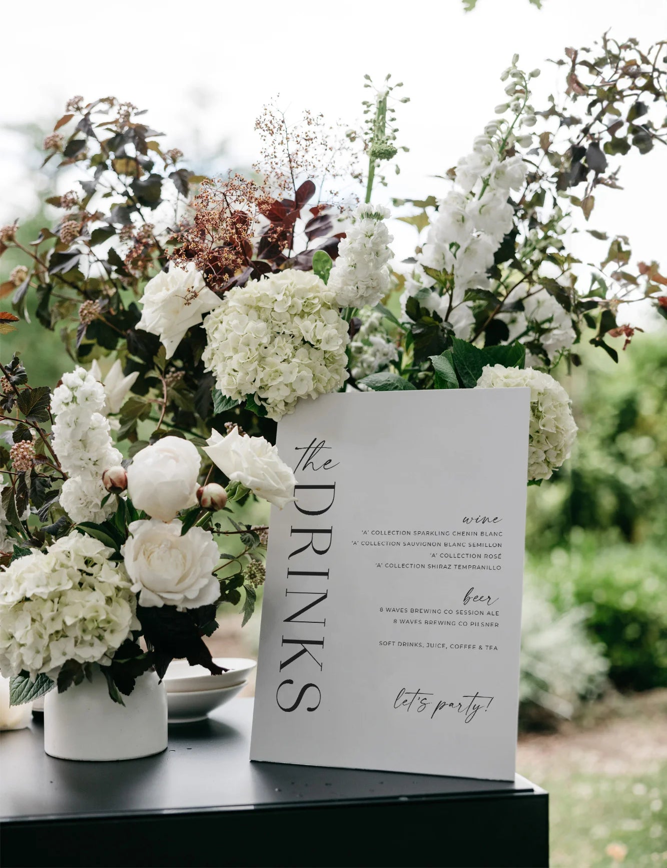 Modern acrylic bar sign featuring a stylish drinks menu with clean typography, surrounded by lush white floral arrangements on a black bar surface at an outdoor venue.

