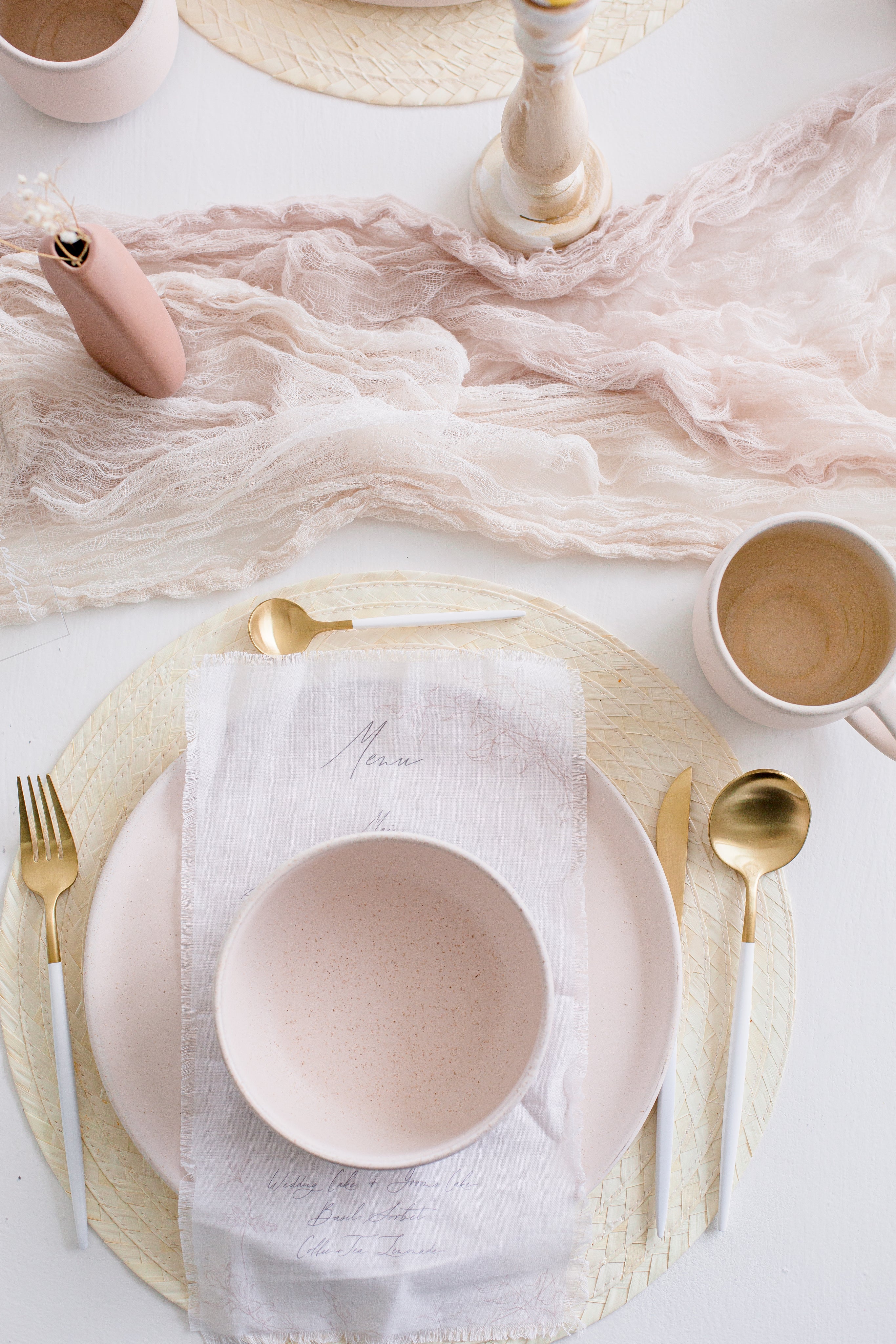 Minimalist wedding table setting with soft blush tones, featuring a linnen menu, a pastel bowl, gold cutlery, and a delicate fabric table runner