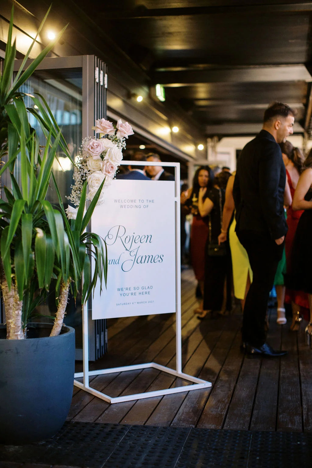 A white wedding welcome sign for "Rojen and James" is displayed on a modern white metal stand. The sign is adorned with soft pink and white roses, placed indoors at a wedding venue. Guests are seen mingling in the background, dressed for the occasion. A tall potted plant sits to the left, adding greenery to the elegant decor.